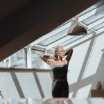 A woman standing in a sunlit attic, arms behind her head, exuding tranquility.