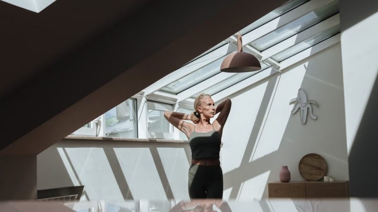 A woman standing in a sunlit attic, arms behind her head, exuding tranquility.