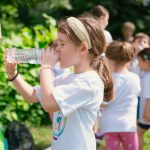 Young girl drinking water during sunny outdoor activity with friends in a park setting.